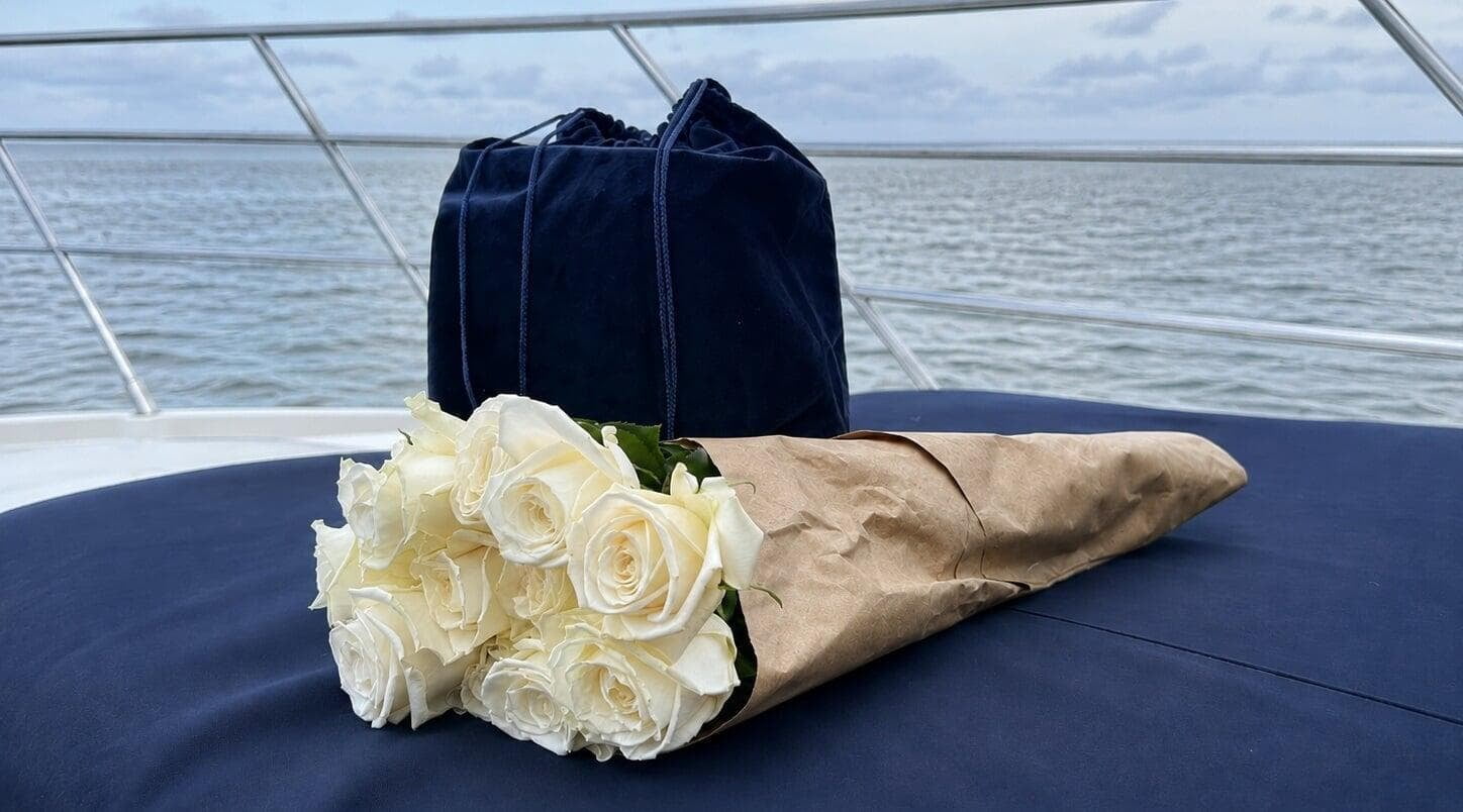 Navy velvet urn bag and a bouquet of white roses resting on a boat cushion before an ash scattering at sea in Miami